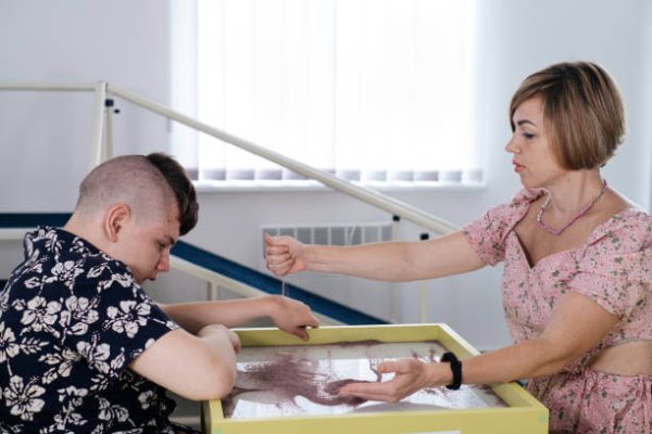 Children with disability getting sensory activity Sand tray, sand table for animation, cerebral palsy boy playing calming game, training fine motor skills. Rehabilitation center with therapist, mother